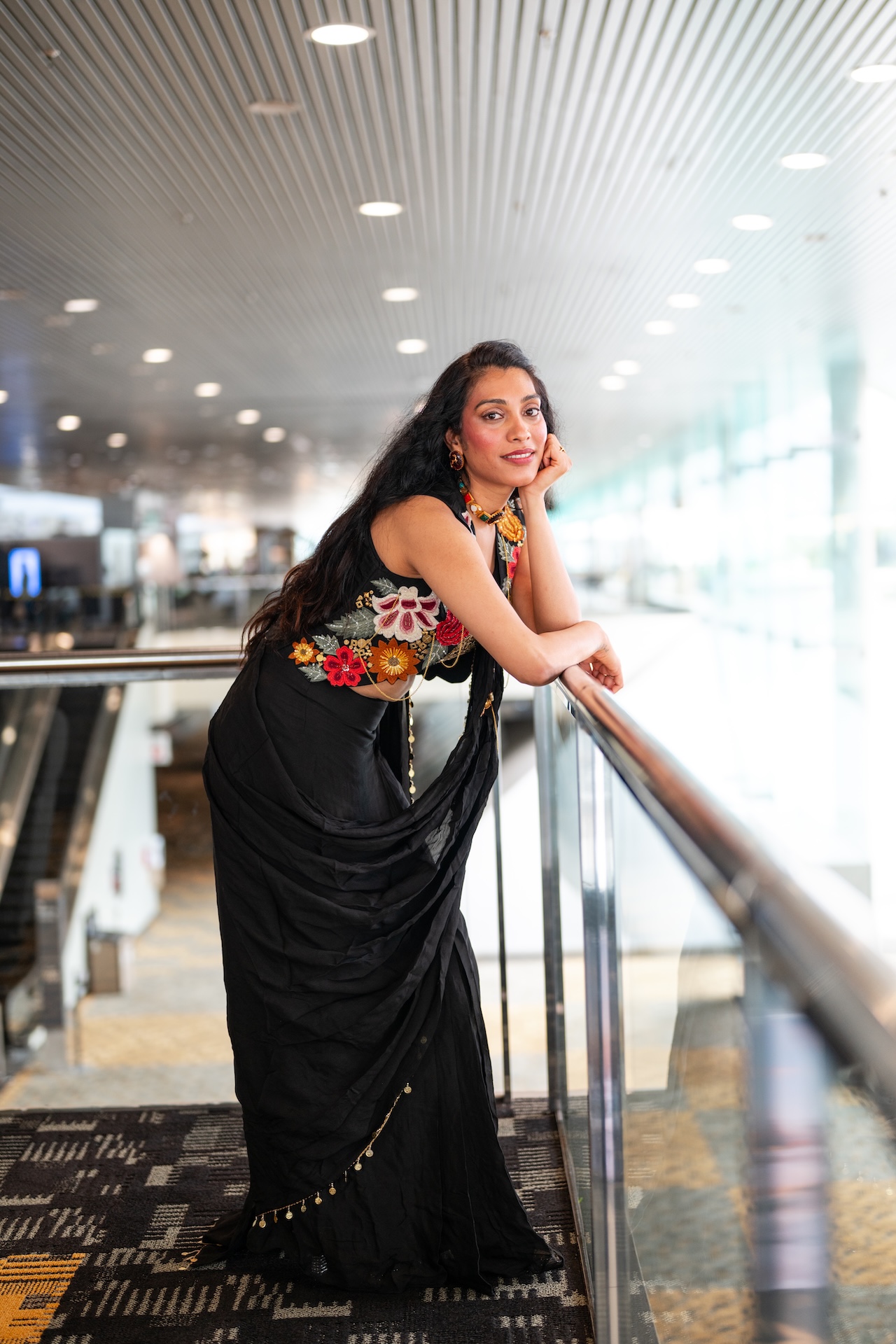 Black embroidered saree leaning on a glass railing in an airy terminal — Vetri Frames, Virginia